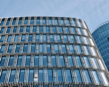 The image features a modern glass office building with a facade of vertical and horizontal metal slats. The building reflects the blue sky, creating a sleek urban look.