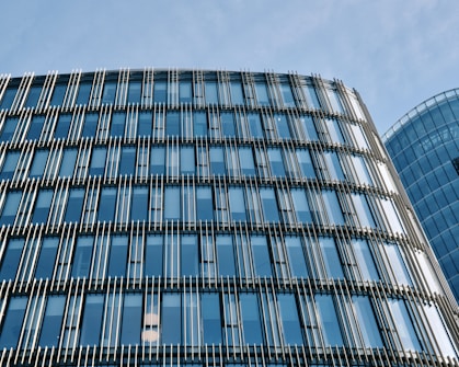 The image features a modern glass office building with a facade of vertical and horizontal metal slats. The building reflects the blue sky, creating a sleek urban look.