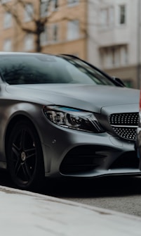 A sleek, modern silver car is parked on the street, partially obscured, with a focus on the front section including the headlights and grille. The background is a blurred urban setting with buildings and trees out of focus, conveying depth and a city environment.