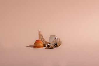 A quail egg is broken open, with the pieces lying on a smooth, beige surface. One of the egg pieces is half of the shell, displaying its speckled brown and white exterior, alongside the clear, shiny inside. Next to it, a caramel-colored yolk is partially intact, providing an abstract yet natural focal point.