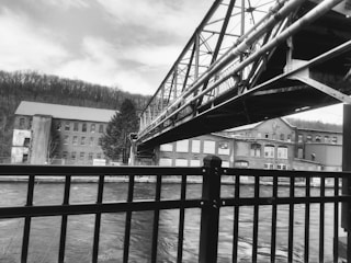 A black and white image depicting an industrial scene with a bridge crossing over a river. To the side, old brick buildings with many windows evoke a sense of historic architecture. The foreground features a metal railing, while hills with trees are visible in the background.