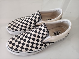 A pair of slip-on shoes with a classic black and white checkerboard pattern on the fabric. The shoes have white rubber soles and visible branding on the inner sole and side tags. They are positioned side by side on a light-colored surface.