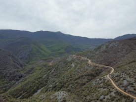A winding dirt path cuts through a rugged mountain landscape covered with dense green vegetation and patches of rocky terrain. Misty clouds hover over the peaks in the background, creating a serene and unspoiled natural setting.
