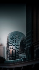 A modern architectural structure with Arabic calligraphy covering its reflective silver surface, set against the backdrop of tall buildings and a dusky sky. The architecture blends contemporary design with traditional art forms.