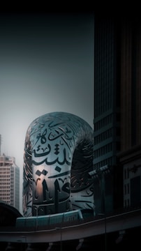 A modern architectural structure with Arabic calligraphy covering its reflective silver surface, set against the backdrop of tall buildings and a dusky sky. The architecture blends contemporary design with traditional art forms.