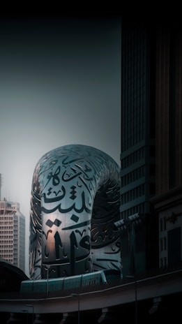 A modern architectural structure with Arabic calligraphy covering its reflective silver surface, set against the backdrop of tall buildings and a dusky sky. The architecture blends contemporary design with traditional art forms.