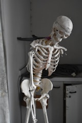 A life-size anatomical skeleton model stands in a room with a slightly tilted posture. The skeleton appears weathered and is positioned next to a cabinet with a coiled wire on top. The lighting is dim, casting shadows that give the scene a somber appearance.