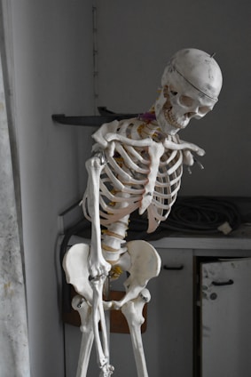 A life-size anatomical skeleton model stands in a room with a slightly tilted posture. The skeleton appears weathered and is positioned next to a cabinet with a coiled wire on top. The lighting is dim, casting shadows that give the scene a somber appearance.
