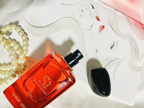 A red perfume bottle labeled 'Giorgio Armani Sì Passione éclat' is placed on a white surface next to a strand of pearls. In the background, a sophisticated sketch of a woman's face with bold red lipstick adds an artistic touch.