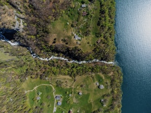 An aerial view of a lush landscape with several houses scattered across green fields and surrounded by trees. A narrow waterway runs through the middle, dividing the land and flowing into a larger body of water on the right.