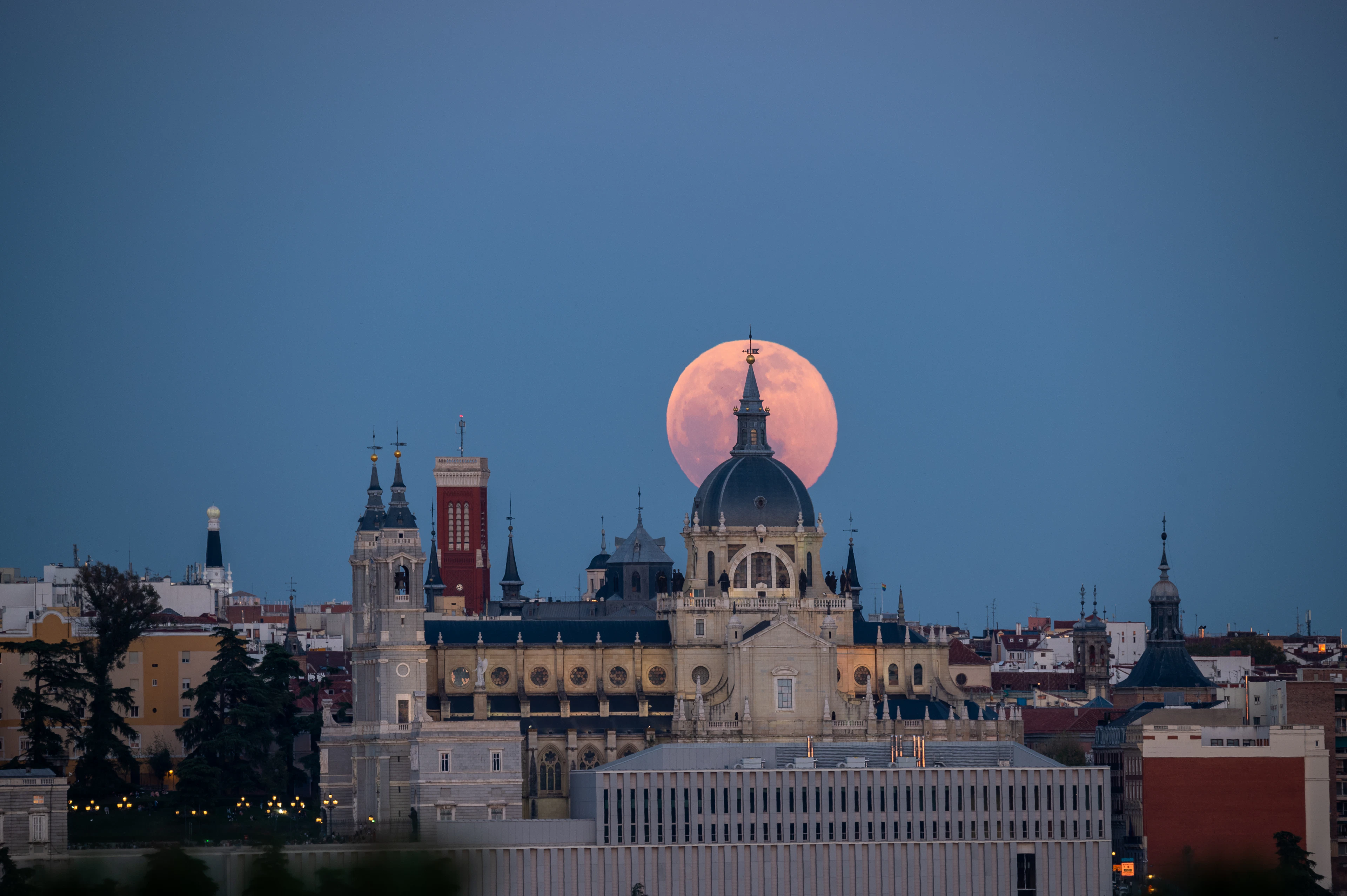 A full moon rising pink behind the domed silhouette of the Almudena Cathedral at blue hour, seen from the west across the rooftops of Madrid.