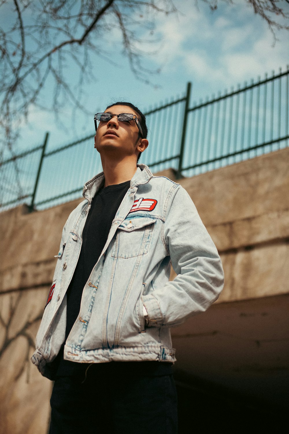 a man wearing a jean jacket and sunglasses standing in front of a bridge