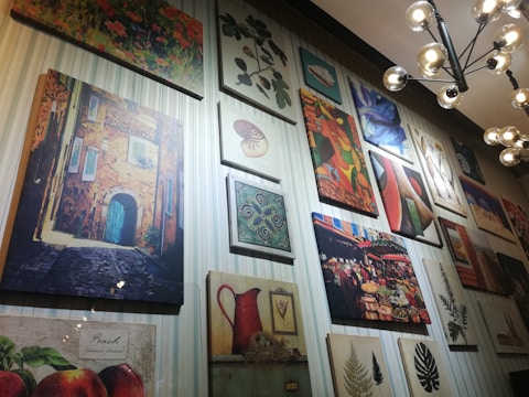 A wall gallery displaying various framed artworks, including a painting of a rustic building with a blue door, a vibrant abstract painting, a botanical print, and a market scene. The lighting is provided by a modern, spherical chandelier.