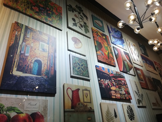 A wall gallery displaying various framed artworks, including a painting of a rustic building with a blue door, a vibrant abstract painting, a botanical print, and a market scene. The lighting is provided by a modern, spherical chandelier.