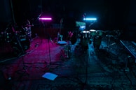 A dimly lit music studio with various instruments including drums, guitars, and a keyboard. Two neon lights, one pink and one blue, cast colorful glows on the instruments and floor. Microphones on stands are scattered around, and everything rests on a patterned rug.