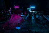 A dimly lit music studio with various instruments including drums, guitars, and a keyboard. Two neon lights, one pink and one blue, cast colorful glows on the instruments and floor. Microphones on stands are scattered around, and everything rests on a patterned rug.