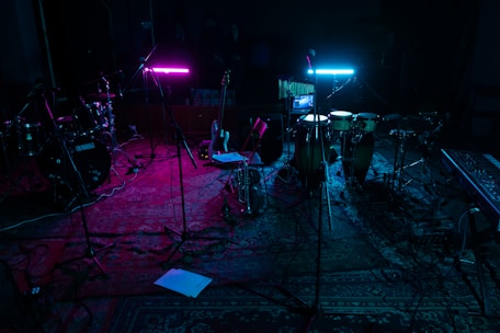 A dimly lit music studio with various instruments including drums, guitars, and a keyboard. Two neon lights, one pink and one blue, cast colorful glows on the instruments and floor. Microphones on stands are scattered around, and everything rests on a patterned rug.