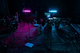 A dimly lit music studio with various instruments including drums, guitars, and a keyboard. Two neon lights, one pink and one blue, cast colorful glows on the instruments and floor. Microphones on stands are scattered around, and everything rests on a patterned rug.