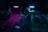 A dimly lit music studio with various instruments including drums, guitars, and a keyboard. Two neon lights, one pink and one blue, cast colorful glows on the instruments and floor. Microphones on stands are scattered around, and everything rests on a patterned rug.
