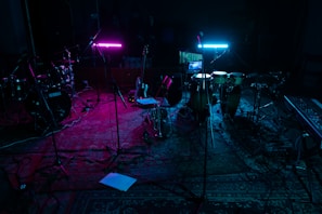 A dimly lit music studio with various instruments including drums, guitars, and a keyboard. Two neon lights, one pink and one blue, cast colorful glows on the instruments and floor. Microphones on stands are scattered around, and everything rests on a patterned rug.