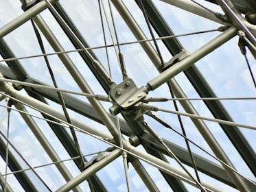 A complex arrangement of metal rods and cables forms a geometric pattern against a backdrop of large, glass panels. The structure appears industrial and showcases precision engineering, with bolts and fittings connecting the various components.