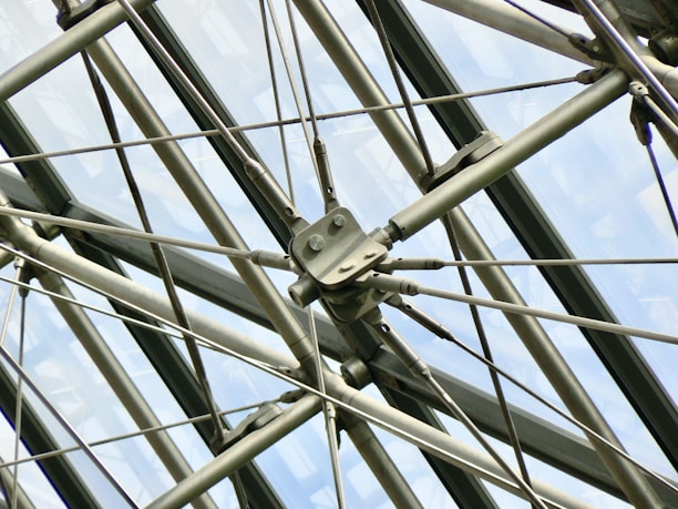 A complex arrangement of metal rods and cables forms a geometric pattern against a backdrop of large, glass panels. The structure appears industrial and showcases precision engineering, with bolts and fittings connecting the various components.