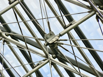 A complex arrangement of metal rods and cables forms a geometric pattern against a backdrop of large, glass panels. The structure appears industrial and showcases precision engineering, with bolts and fittings connecting the various components.
