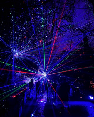 A mesmerizing night scene featuring vibrant laser lights in a forested area. The lasers create patterns of blue, green, and red beams radiating outwards, illuminating the silhouettes of people walking through the space. The dense trees are visible in the background, adding depth and mystery to the glowing atmosphere.