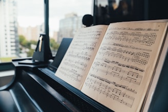 A close-up view of an open music book placed on a piano, with sheet music clearly visible. In the background, there is a blurred view of buildings and trees through a large window, giving a sense of being in a high-rise urban space.