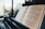 A close-up view of an open music book placed on a piano, with sheet music clearly visible. In the background, there is a blurred view of buildings and trees through a large window, giving a sense of being in a high-rise urban space.