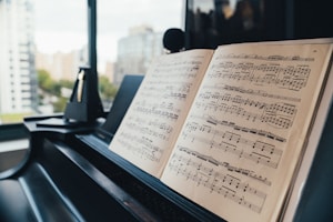 A close-up view of an open music book placed on a piano, with sheet music clearly visible. In the background, there is a blurred view of buildings and trees through a large window, giving a sense of being in a high-rise urban space.