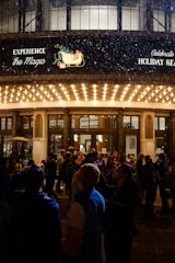 A crowd of people gathered outside a well-lit building entrance during nighttime, possibly for a holiday event. Snow is gently falling, adding to the festive atmosphere. A marquee overhead displays holiday-themed signage and images.