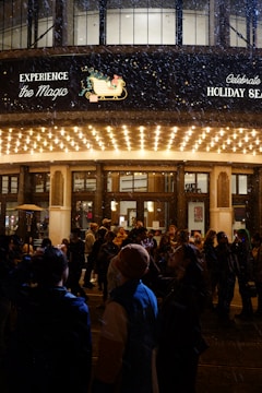 A crowd of people gathered outside a well-lit building entrance during nighttime, possibly for a holiday event. Snow is gently falling, adding to the festive atmosphere. A marquee overhead displays holiday-themed signage and images.