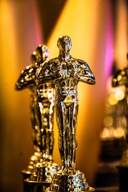 Golden statuettes resembling human figures are positioned closely together. They feature a glossy finish with a prominent base and are set against a softly blurred background with warm lighting.