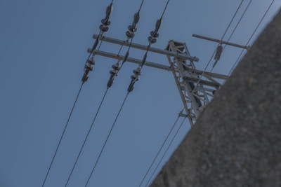 A high-voltage power line structure with insulators and wires extending against a clear blue sky. Part of a concrete structure or building is visible in the bottom right corner.