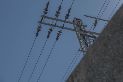 A high-voltage power line structure with insulators and wires extending against a clear blue sky. Part of a concrete structure or building is visible in the bottom right corner.
