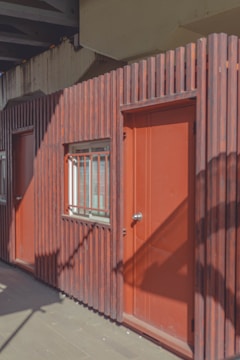A wooden facade featuring vertical slats painted in a warm reddish-brown tone. There are two doors and a window, each with metal hardware, set against the slatted design. The area is lit by sunlight casting shadows of nearby structures.
