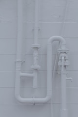 Several industrial pipes are mounted vertically against a smooth, light-colored wall. The pipes are well-organized, creating a geometric pattern with bends and joints. The wall has horizontal lines, adding texture to the background.