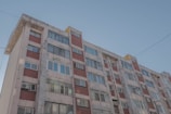 A multi-story residential building with a worn facade. The structure features a mix of red brick and concrete, with numerous windows reflecting daylight. The building shows signs of aging, with visible wear and patches. Electrical wires are seen above the structure under a clear sky.