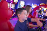 A young boy is enthusiastically playing a driving arcade game. The scene is vibrant with colorful lights and blurred background, indicating a lively arcade environment. Another person, slightly out of focus, is also engaged in a similar activity, adding to the sense of fun and excitement.