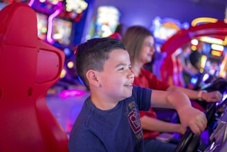 A young boy is enthusiastically playing a driving arcade game. The scene is vibrant with colorful lights and blurred background, indicating a lively arcade environment. Another person, slightly out of focus, is also engaged in a similar activity, adding to the sense of fun and excitement.