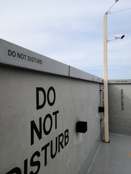 A concrete wall featuring multiple 'Do Not Disturb' signs in bold black letters. A security camera is mounted on a pole extending from the ground, and the sky above is overcast with light gray clouds.