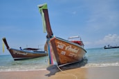 Long-tail boats are docked on a sandy beach with calm ocean waves in the background. The boats have colorful cloths tied to their bows and display Thai script on their sides. The sky is clear with a few scattered clouds.