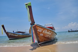 Long-tail boats are docked on a sandy beach with calm ocean waves in the background. The boats have colorful cloths tied to their bows and display Thai script on their sides. The sky is clear with a few scattered clouds.
