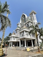 A grand church building with intricate architectural details, featuring tall, pointed arches and decorative elements. Palm trees surround the structure, which is adorned with a large cross at the top. The church has a series of steps leading to the entrance, and a clear blue sky serves as the backdrop.