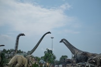 A display of large dinosaur sculptures, some with long necks, set against a backdrop of a cloudy sky. Palm trees and other greenery are visible around the sculptures, along with a central lamppost.