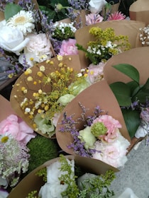 A collection of flower bouquets wrapped in brown paper, featuring a variety of blooms such as white roses, pink peonies, yellow filler flowers, and purple statice. The arrangements are lush with greenery, including leaves and delicate sprigs.