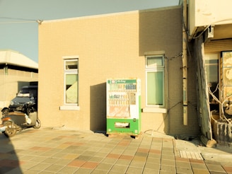 A vending machine is positioned against a brick building wall under direct sunlight. The image displays a paved area in front of the building. A scooter is parked nearby, mostly in the shadow. The scene has a warm, sepia-toned color filter, giving it a nostalgic appearance.