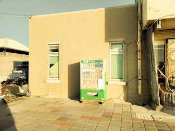 A vending machine is positioned against a brick building wall under direct sunlight. The image displays a paved area in front of the building. A scooter is parked nearby, mostly in the shadow. The scene has a warm, sepia-toned color filter, giving it a nostalgic appearance.