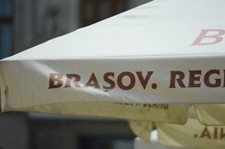 A close-up of a large outdoor umbrella featuring the word 'BRASOV' prominently printed in bold, brown letters on a light-colored fabric. The background consists of a blurred portion of a building and another umbrella underneath.